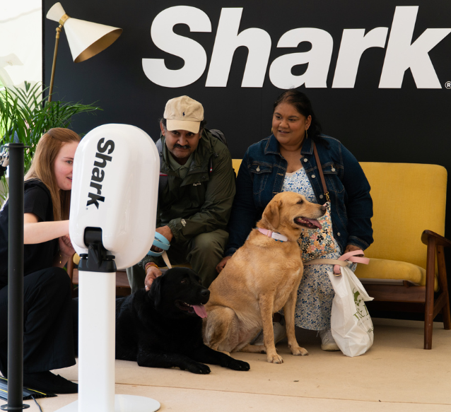 A couple and their two dogs, sat on a sofa, receive a Shark product demo from a lady sat on the floor